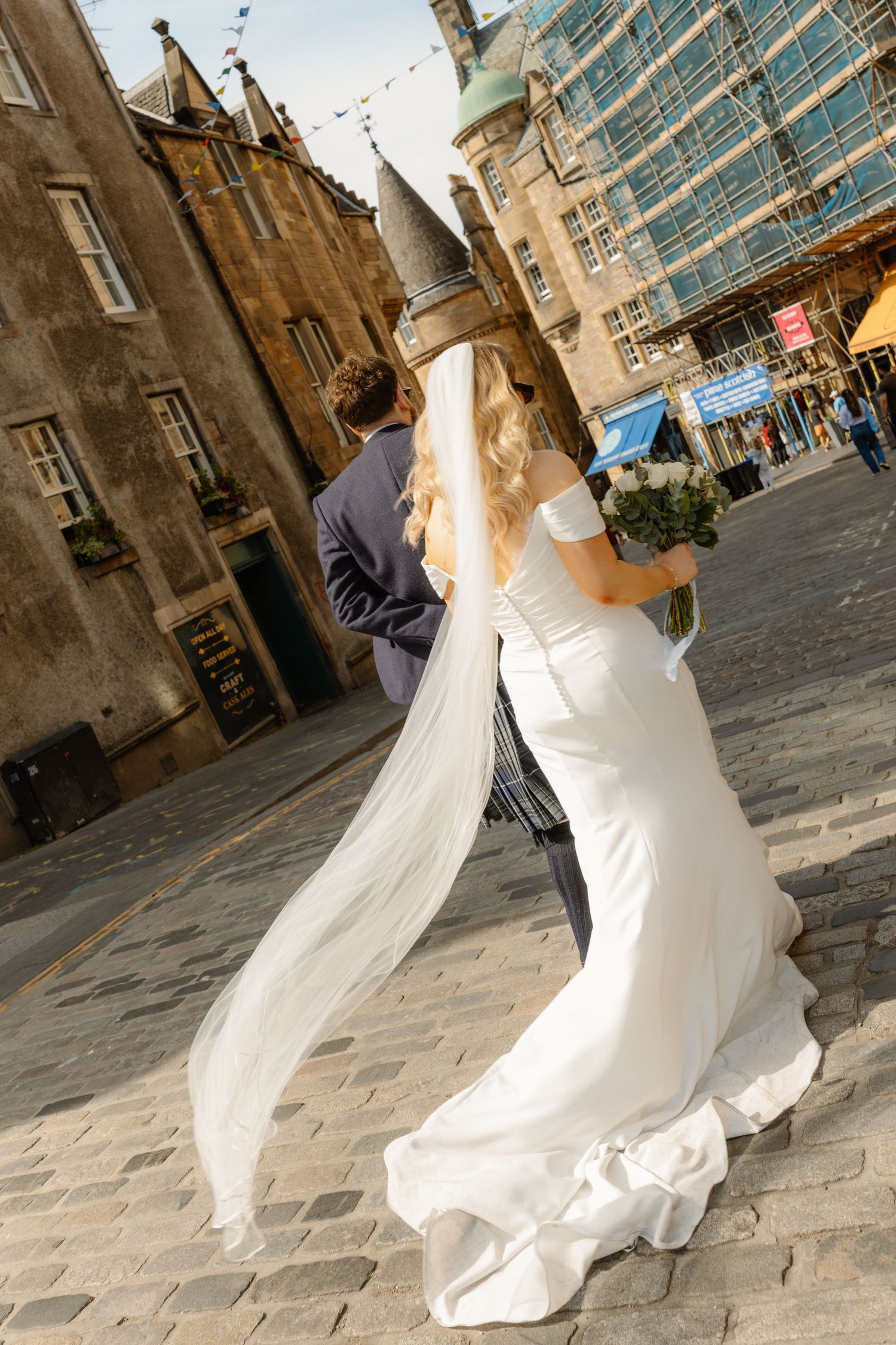 Bride in white gown and veil walks with groom in suit on cobblestone street in historic Edinburgh.