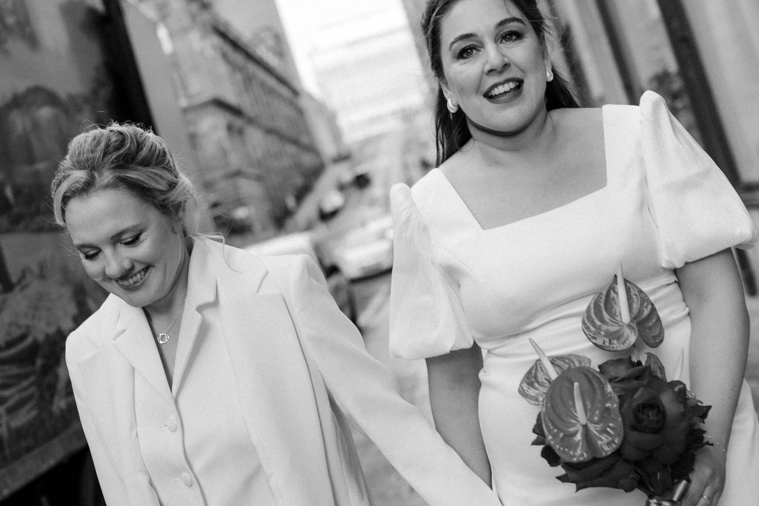 Two brides in white wedding attire walk together, one in a dress holding a bouquet, the other in a chic suit.
