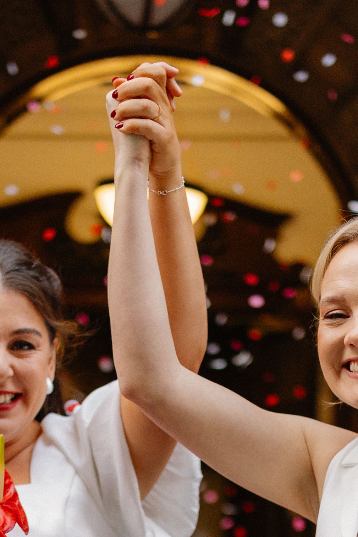 Two brides in white dresses joyfully raise their joined hands amid colorful confetti at their wedding celebration.