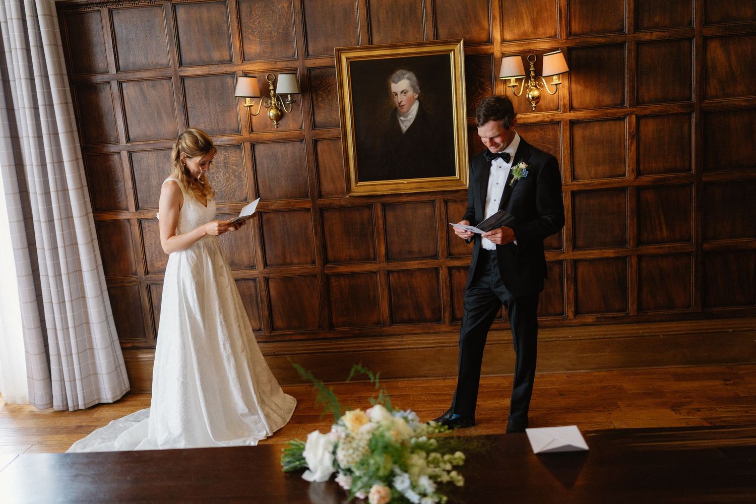 Bride and groom exchanging private vows in wood-paneled room with portrait painting and floral arrangement.