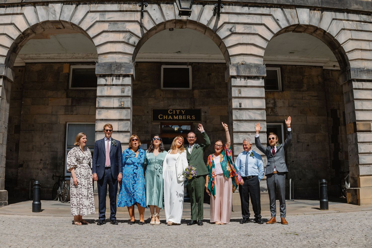 Wedding party celebrating outside Edinburgh City Chambers with raised arms on sunny day.