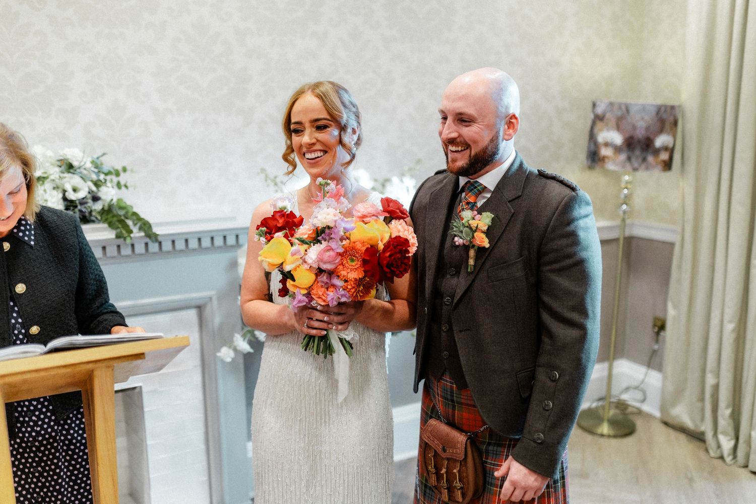 Bride holding colorful bouquet stands with groom in tartan kilt during Scottish wedding ceremony indoors.
