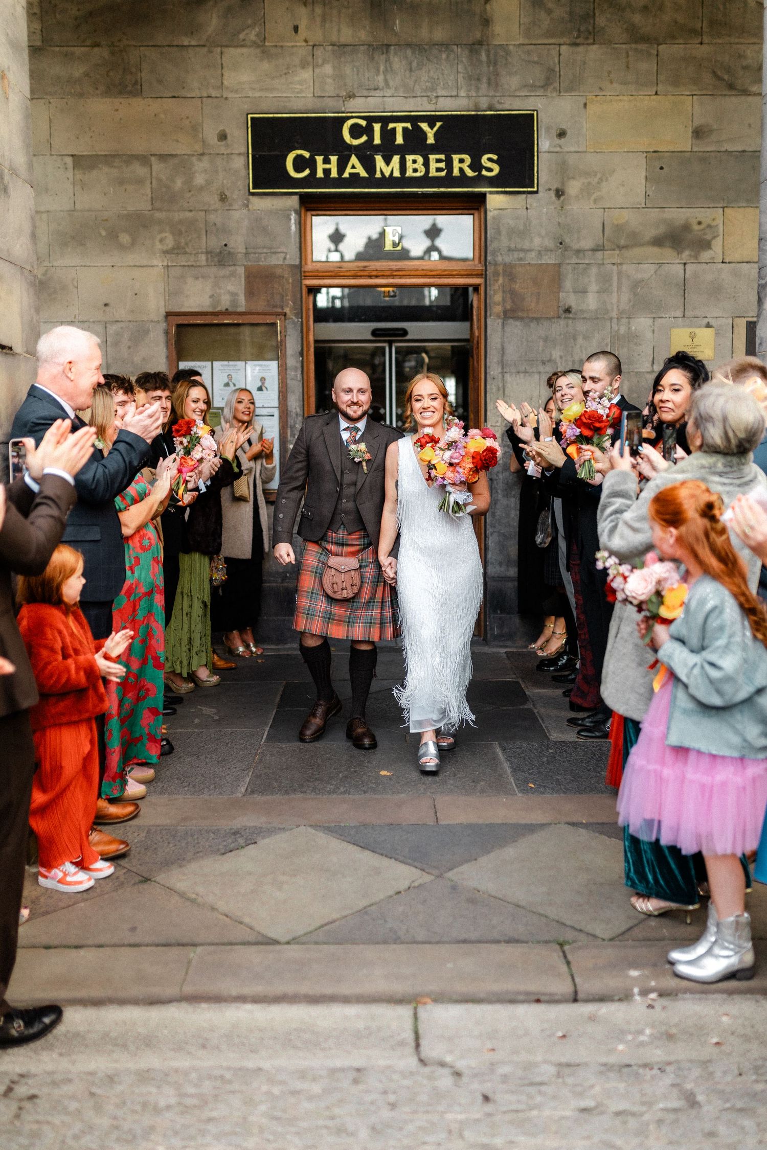 Newlywed couple exits Edinburgh City Chambers through confetti shower as guests cheer and applaud outside.