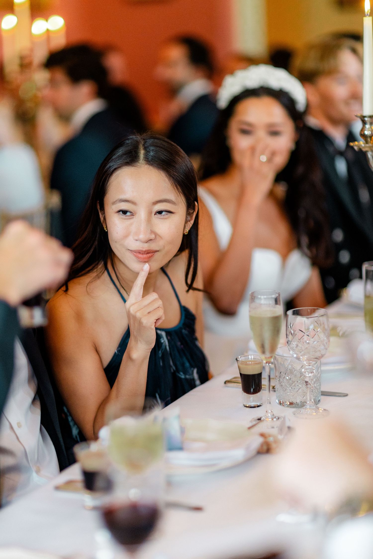 Woman in blue velvet dress gestures at wedding reception table with bride in white dress laughing in background.