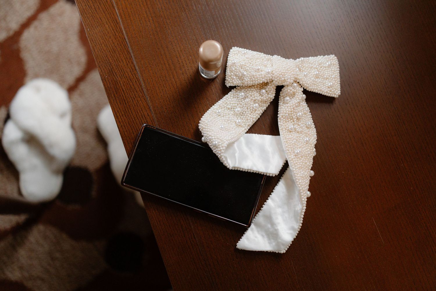 A lace bow hair accessory and smartphone on a wooden table with a small perfume bottle at a wedding.