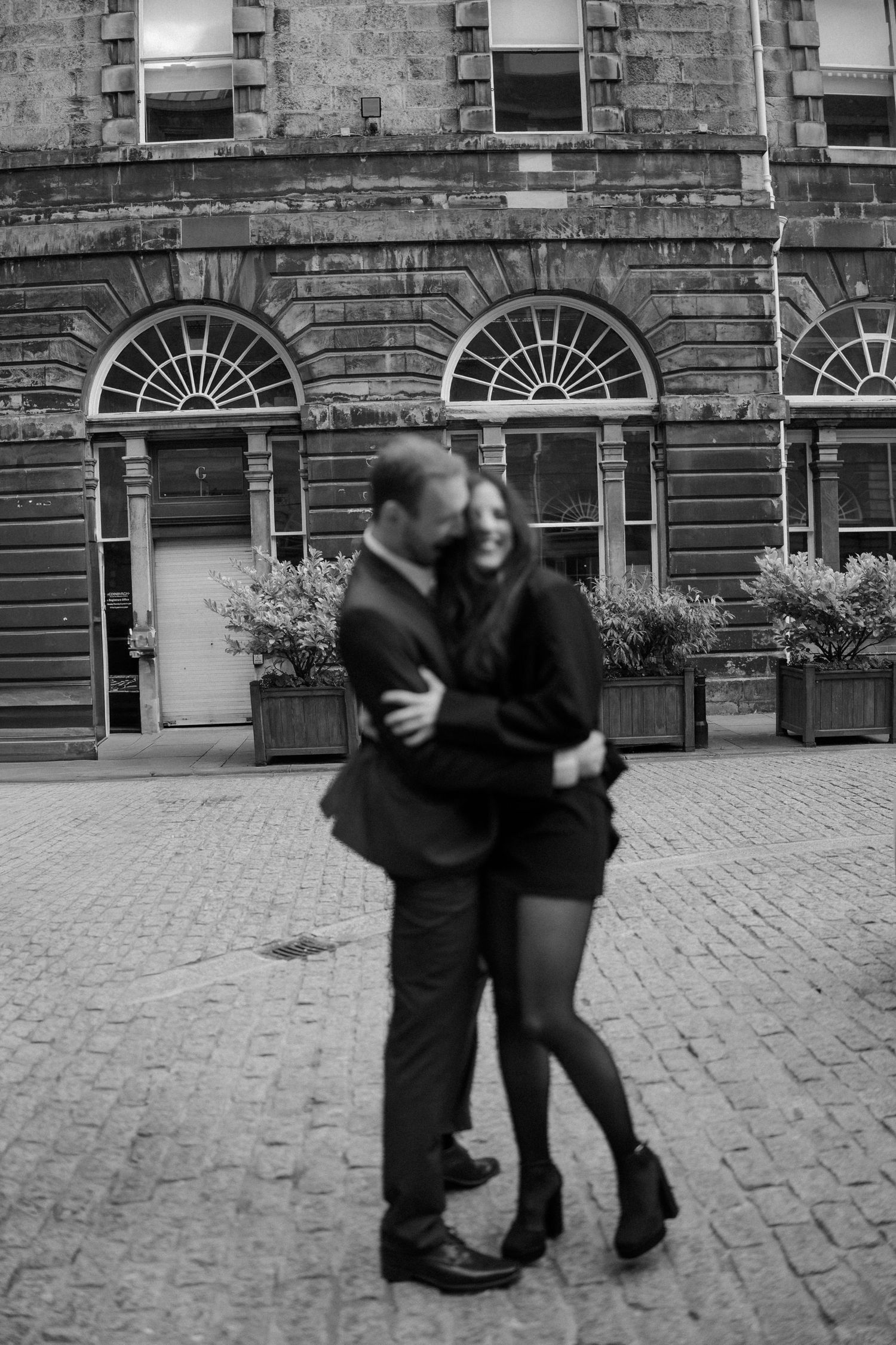 Black and white photo of a romantic couple embracing and laughing on a cobblestone street in front of a Georgian building.