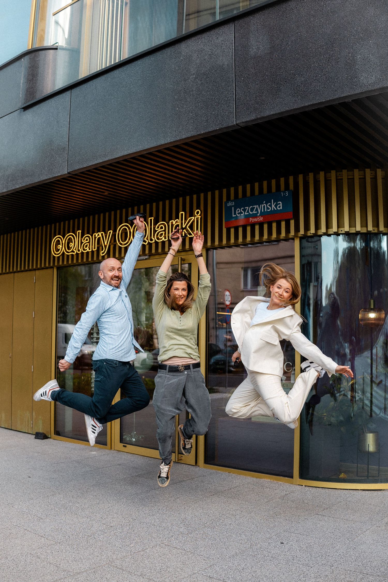 Three people jumping joyfully in front of a storefront with Gallery Opticks signage.