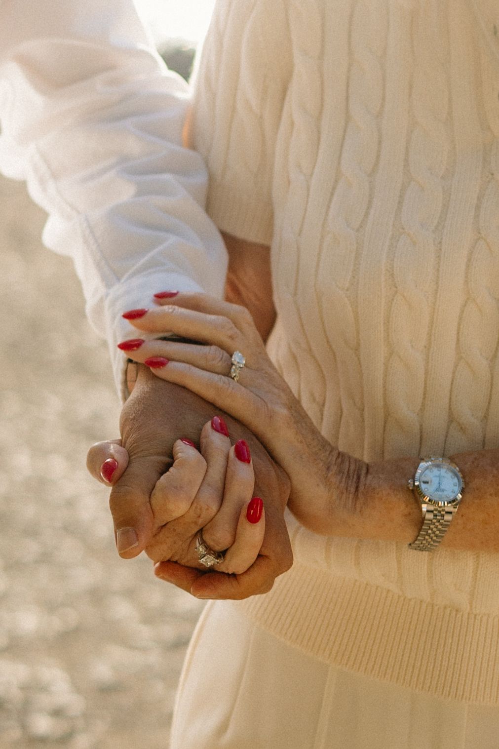 Close-up of hands with engagement ring and red nail polish against a cream sweater.