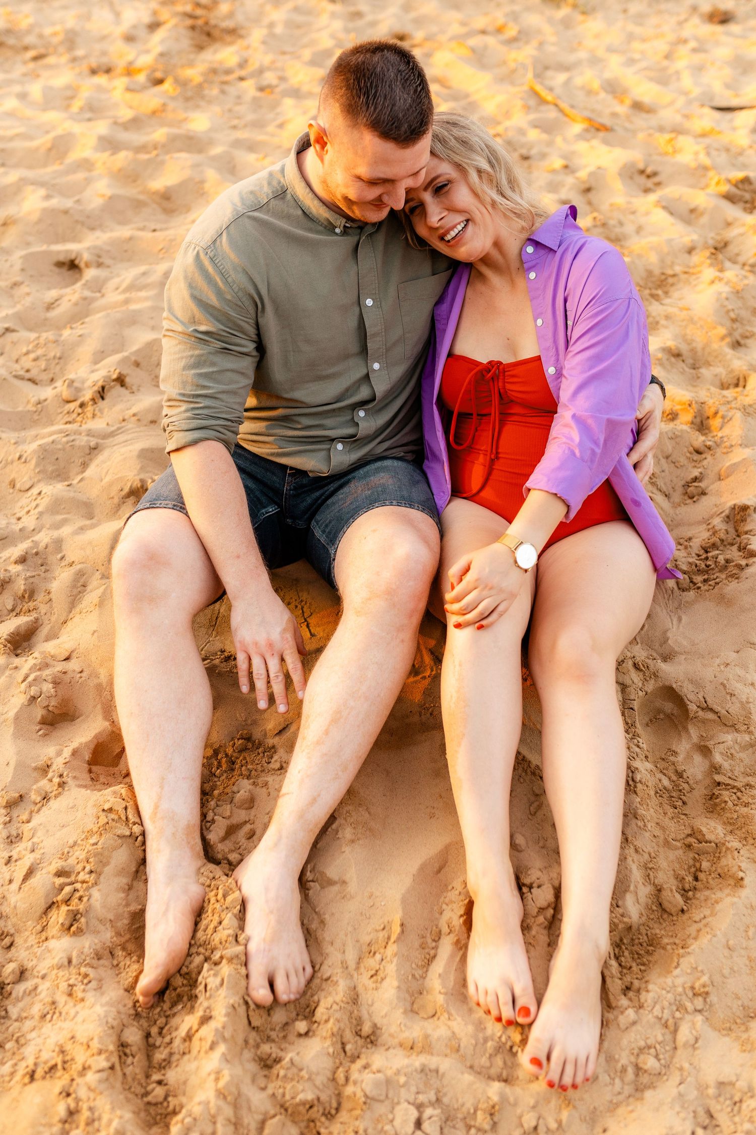 A couple sits together on sandy beach wearing casual summer clothing.