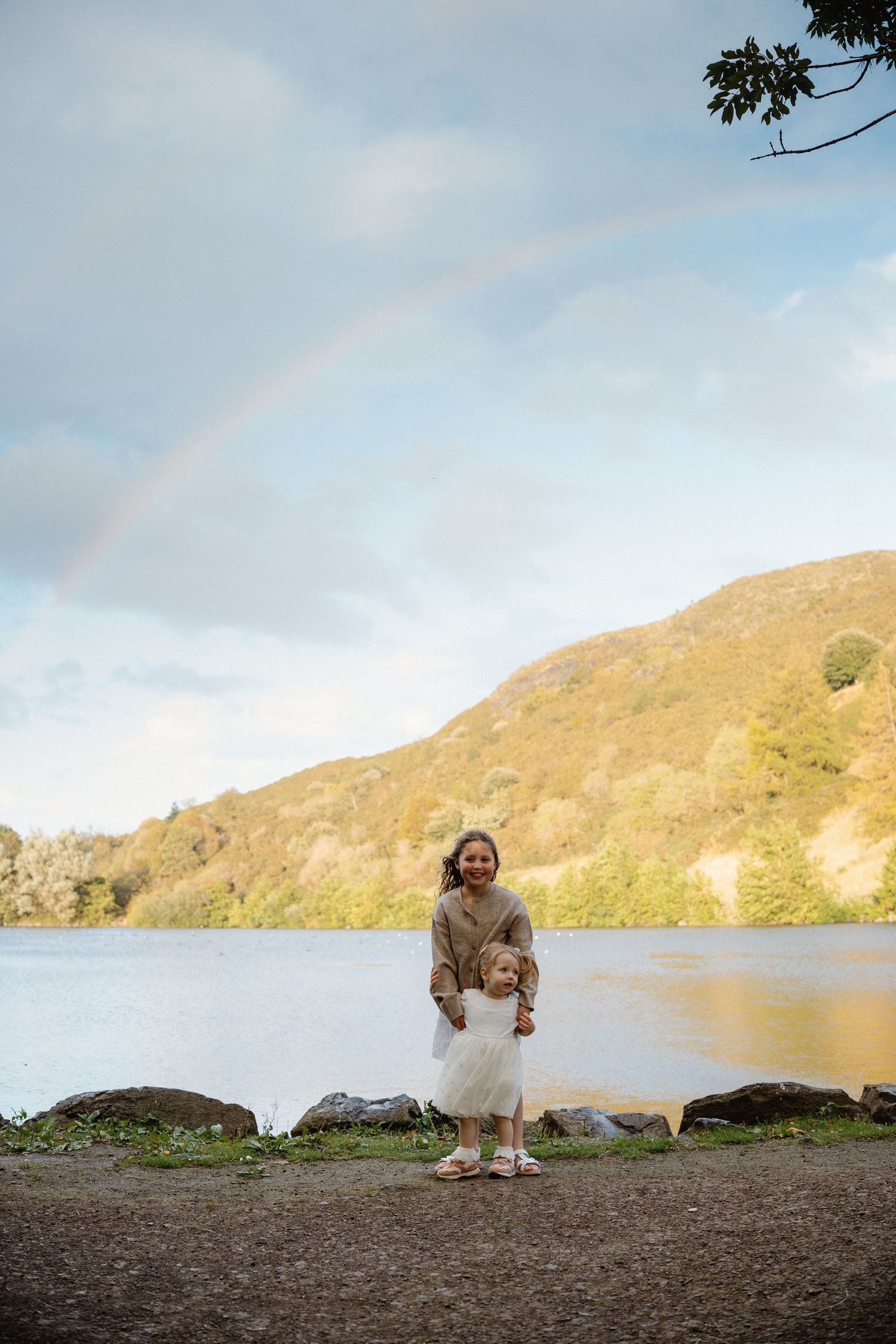 Child in light colored dress plays near scenic lake with mountains in background.