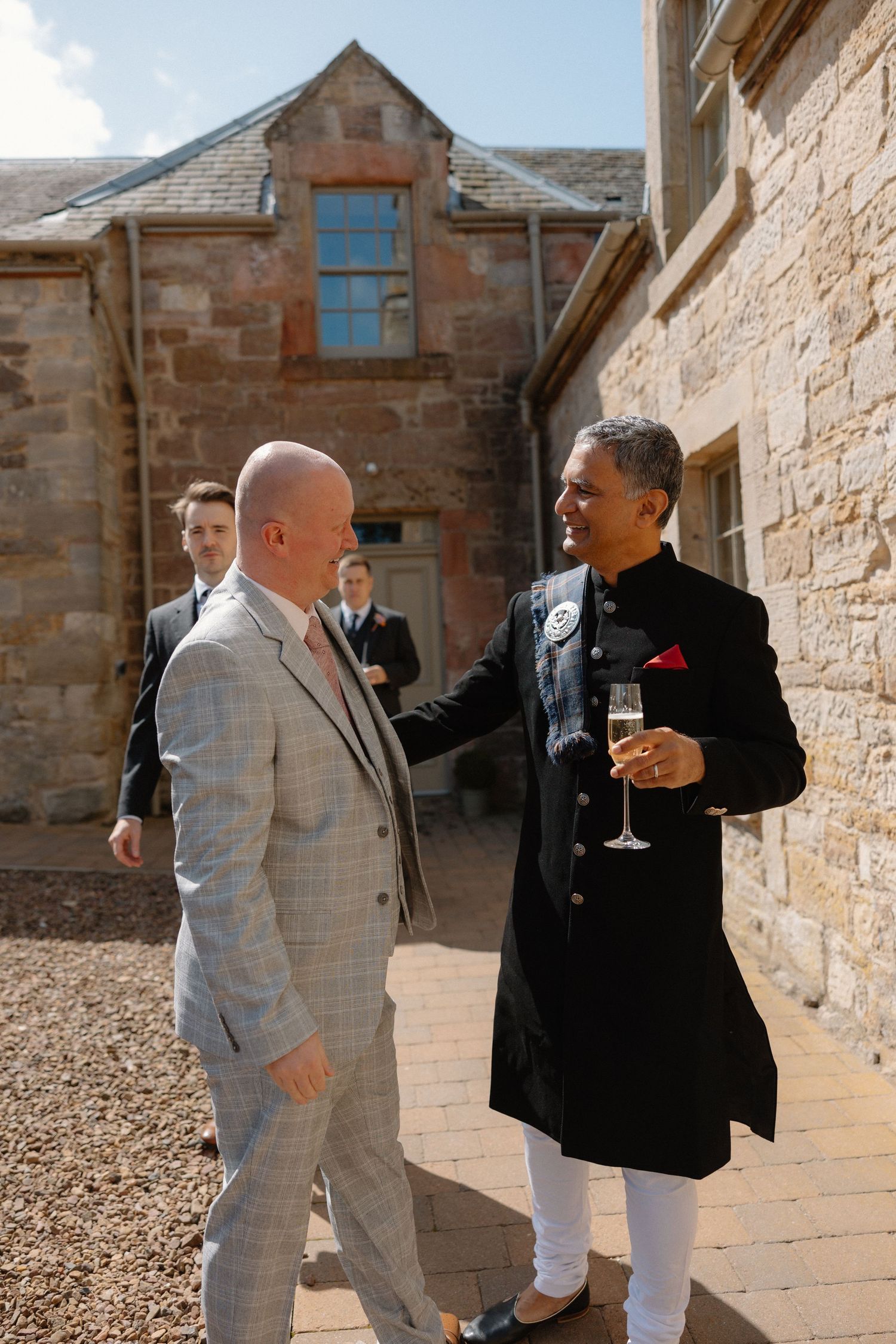 Wedding guests in suits enjoying drinks and conversation outside a stone building.