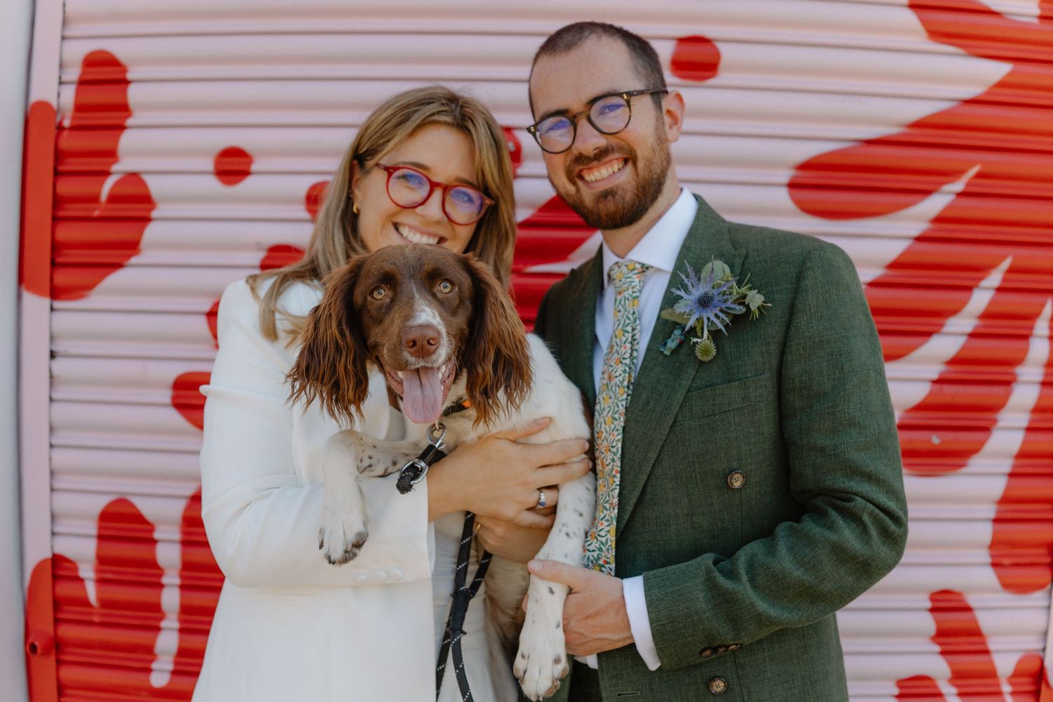 A couple poses with their dog against a white wall with red artistic patterns.
