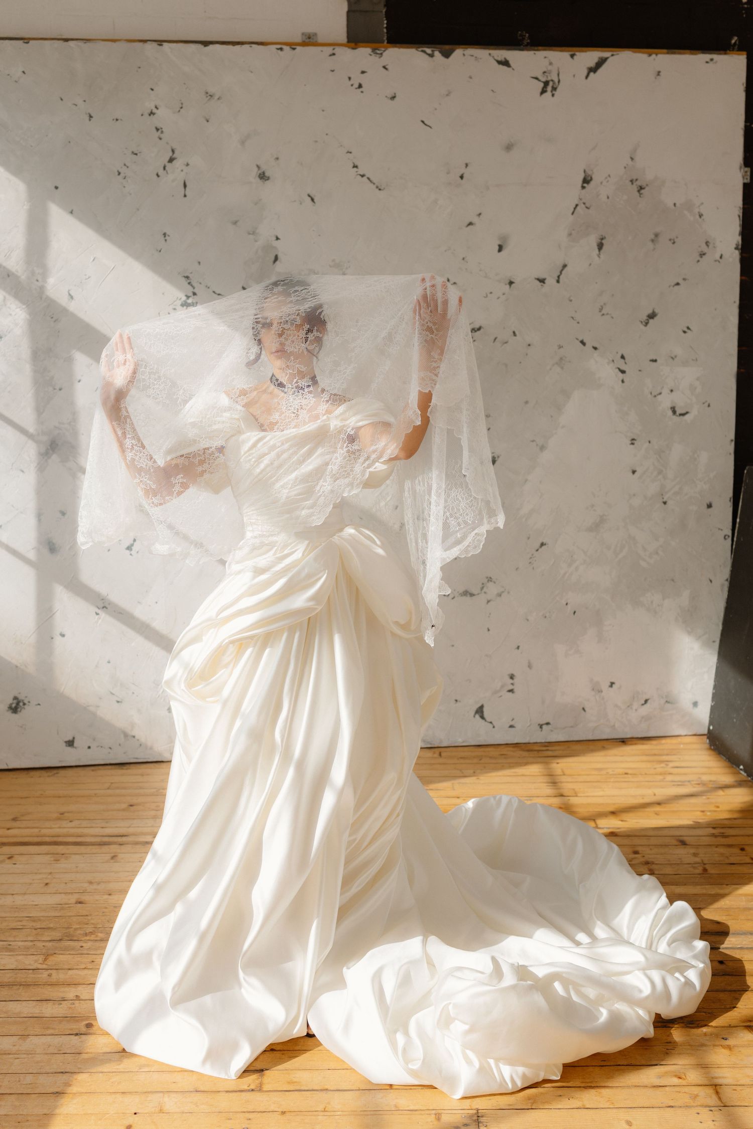 Bride in flowing white satin gown lifting sheer veil, posing in sunlit studio with wooden floors.