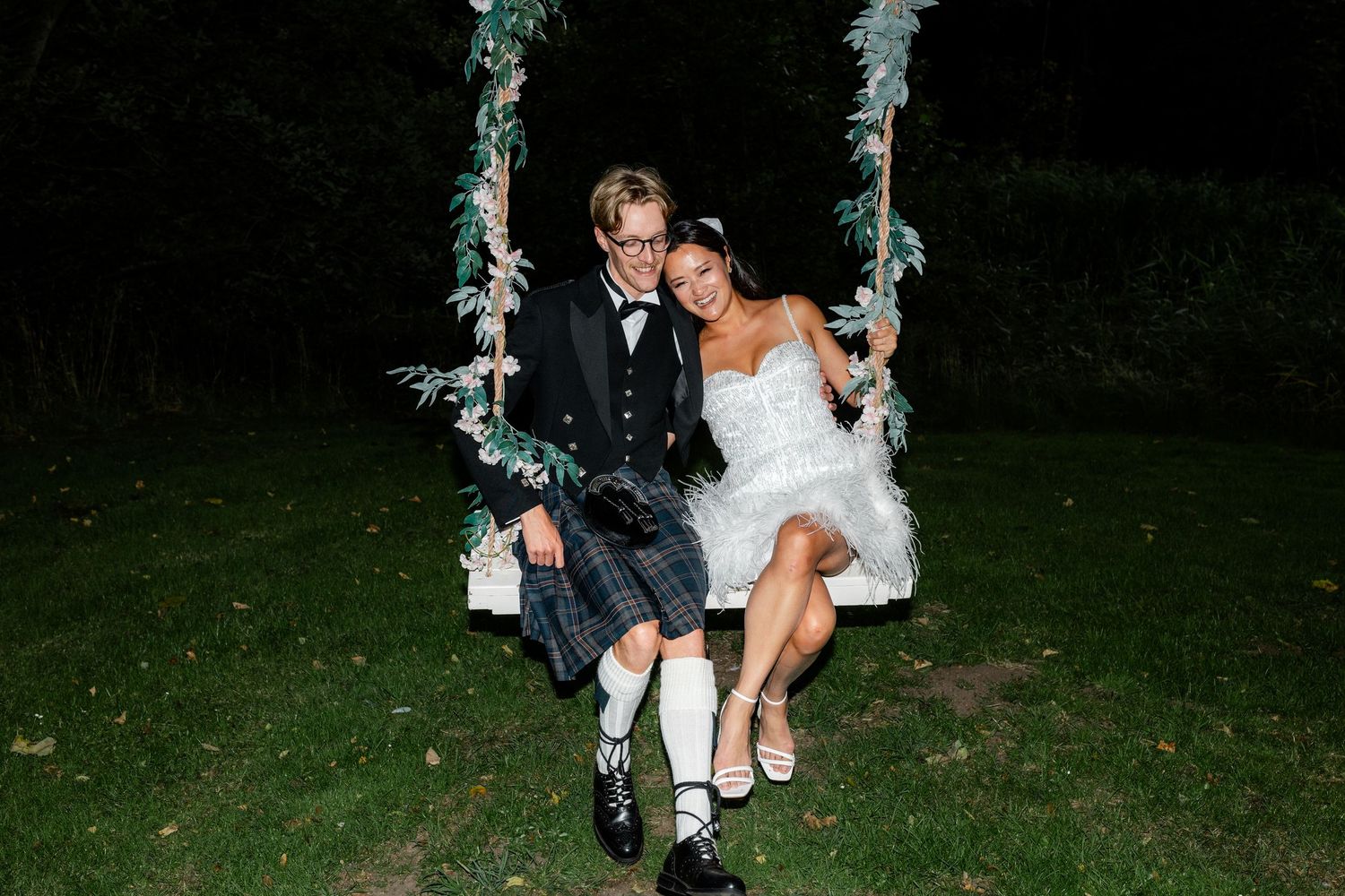 Newlywed couple sitting together on floral swing at night, groom in kilt and bride in short white wedding dress.