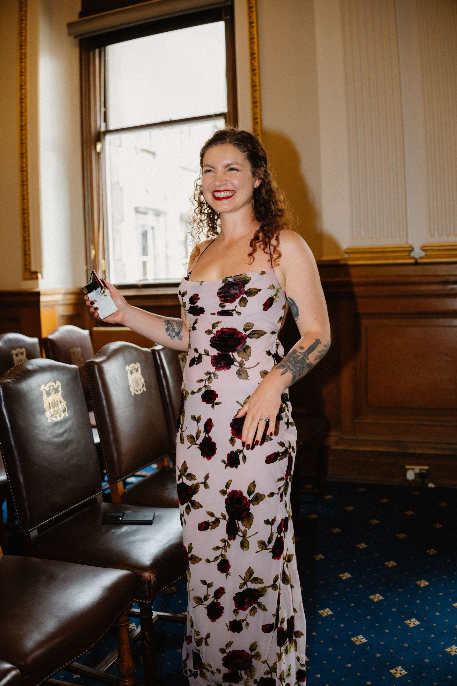 Woman in floral dress smiling at wedding reception holding phone.