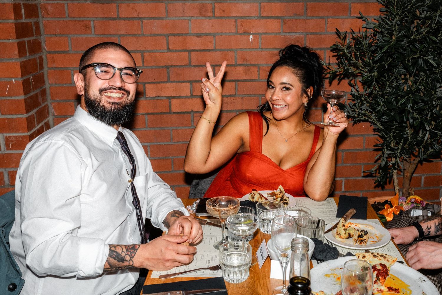 Couple smiling at dinner table with food and drinks, woman in red dress making peace sign at restaurant.