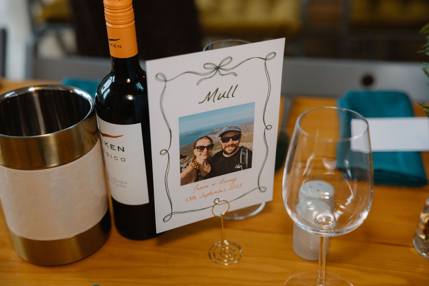 Wedding table setting with wine bottle, personalized photo card labeled Mull, and elegant stemware on wooden table.
