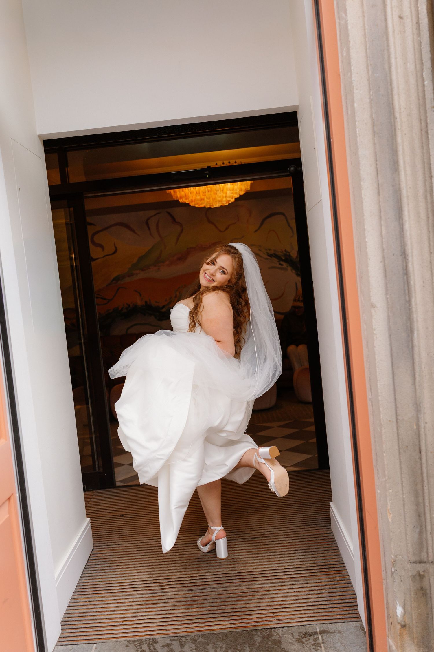 Joyful bride in white gown and veil lifting dress while entering through hotel doorway on wedding day.