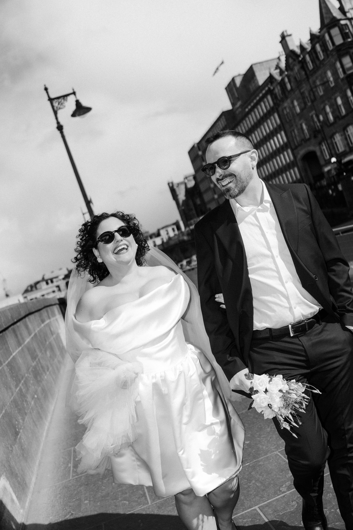 Happy bride and groom in sunglasses walking and laughing on a bridge in black and white photo.