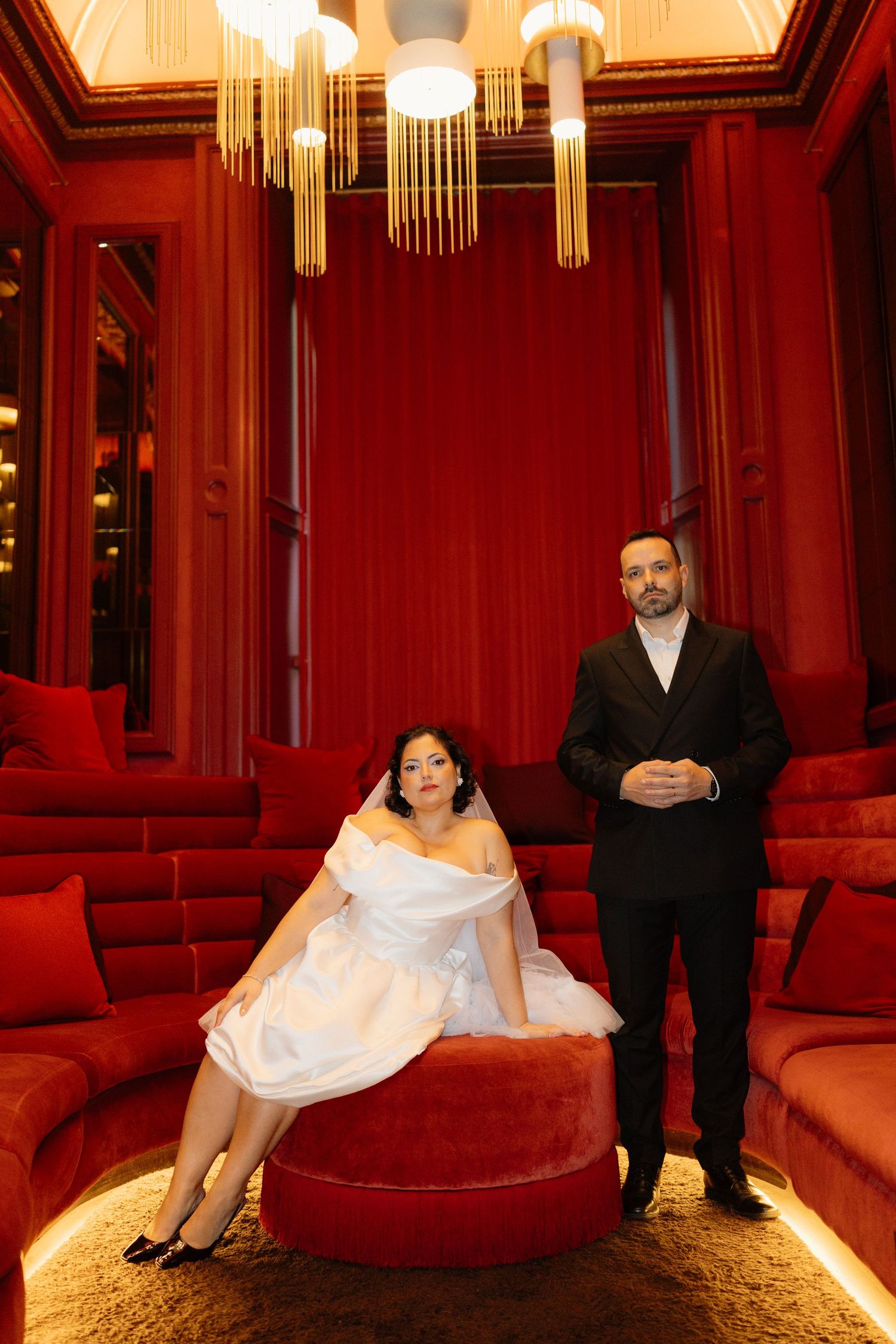 Bride in strapless white mini dress sits on red ottoman beside groom in black suit in opulent red room.