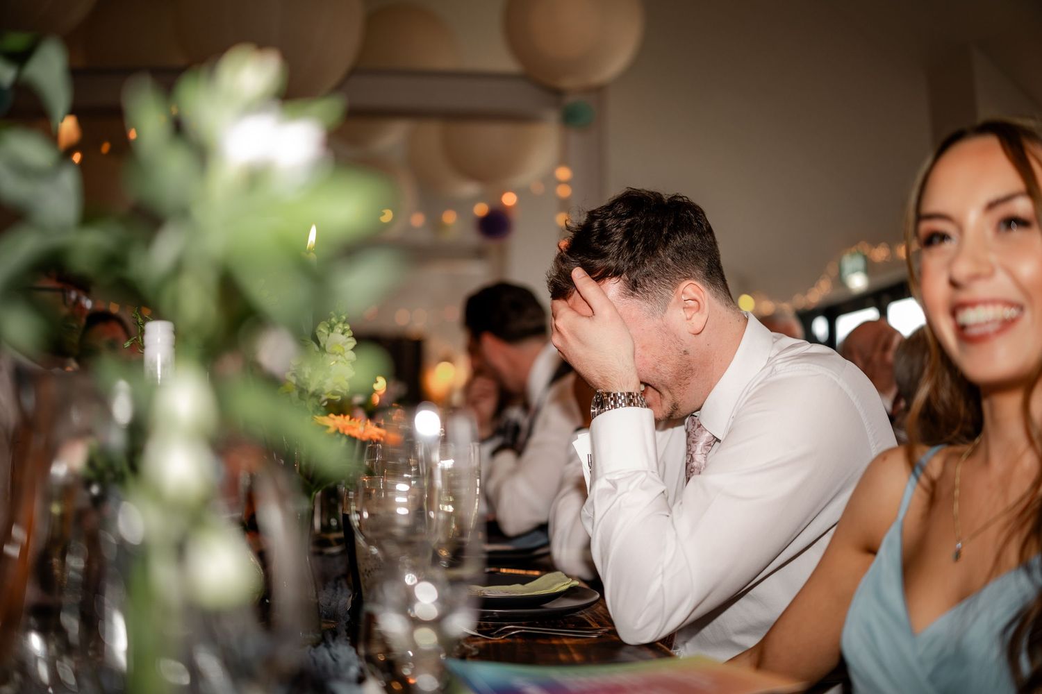 Man covering face with hands laughing at wedding reception dinner table, woman smiling beside him.