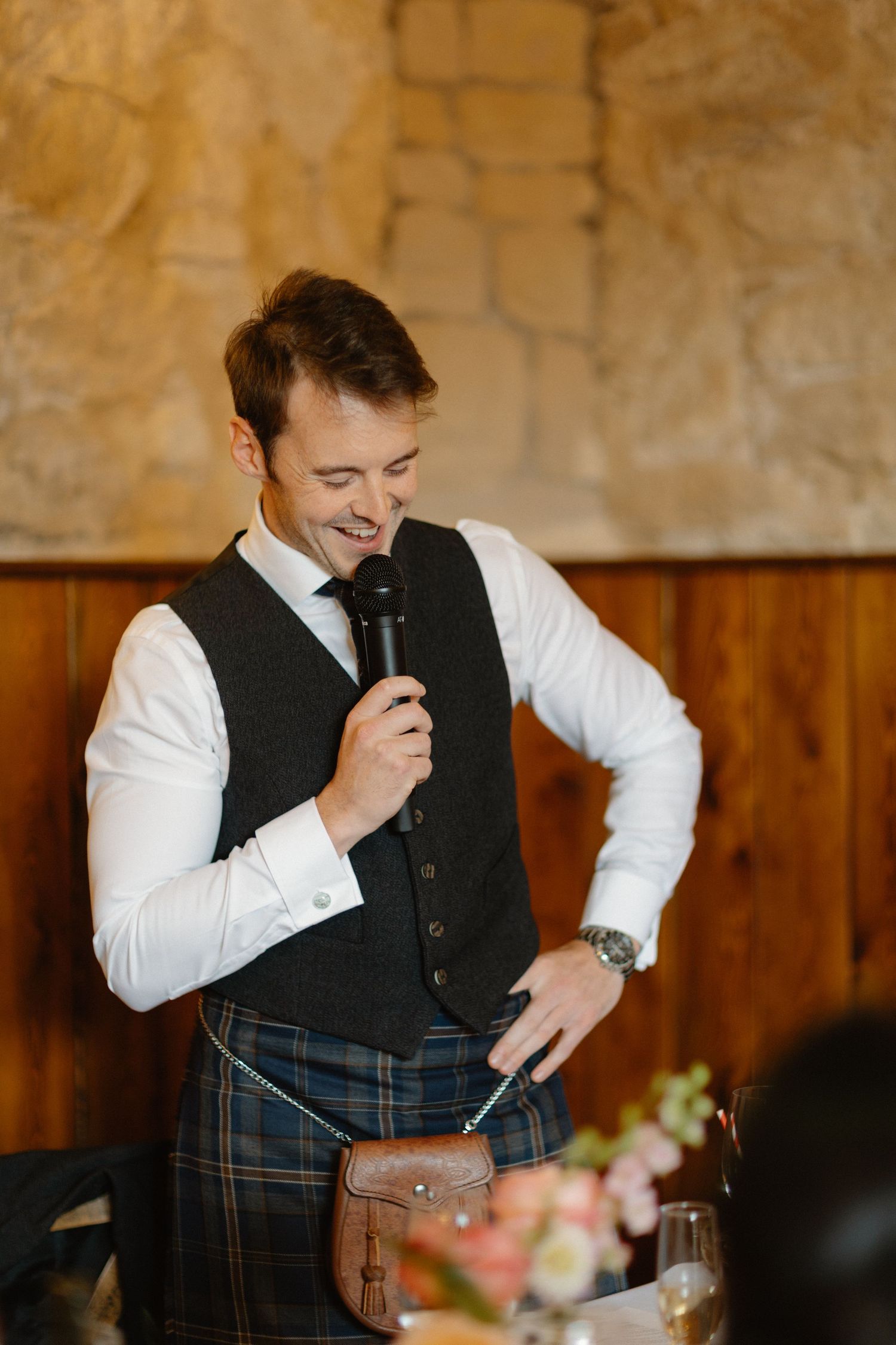 Smiling groom in a tartan kilt and dark waistcoat giving a speech with a microphone at his wedding.