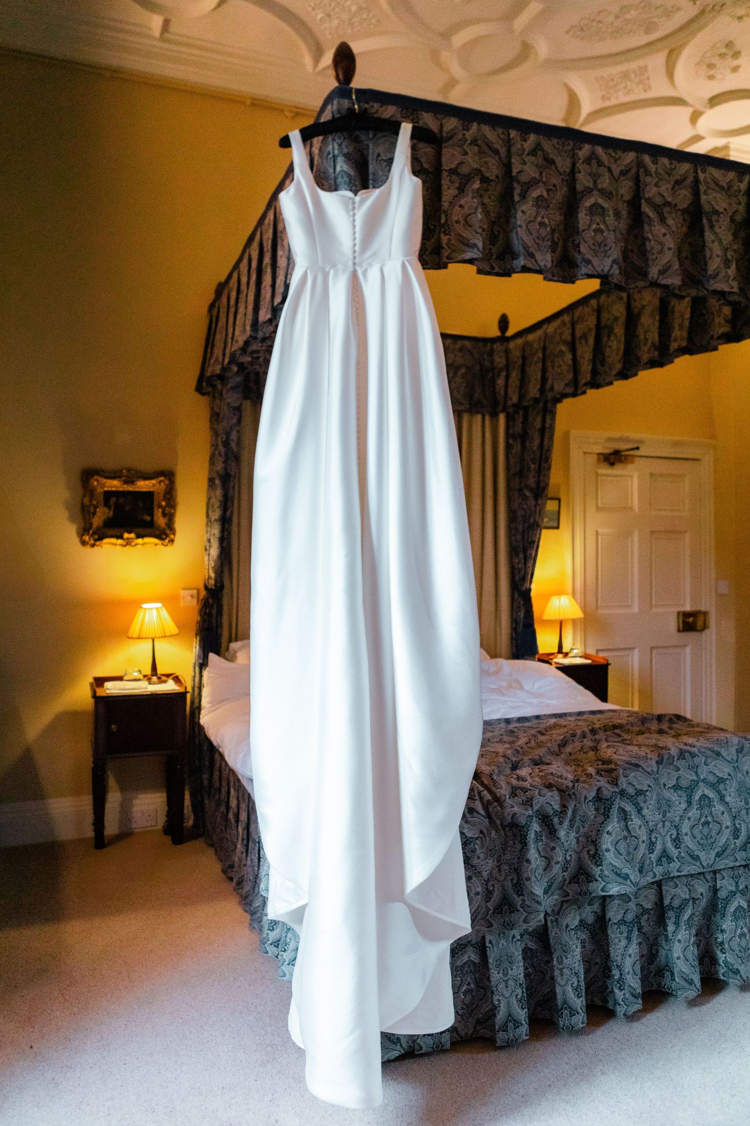 A white wedding dress hangs from a four-poster bed frame in an elegant bedroom with warm lighting and dark bedding.