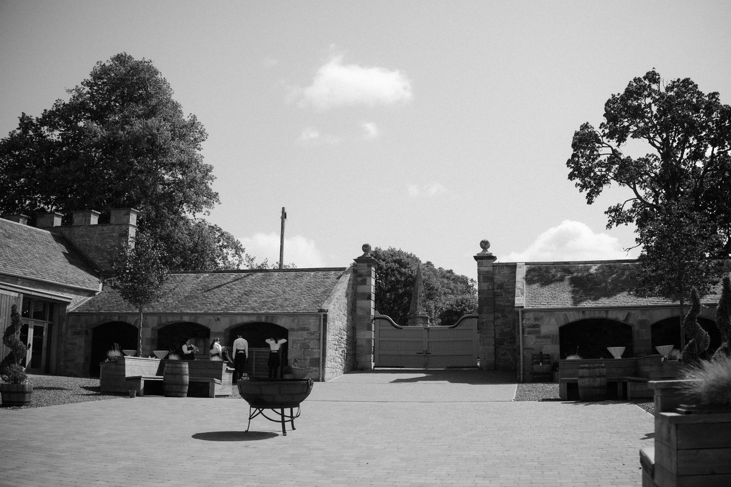 A black and white photograph shows a courtyard with stone buildings and trees against a cloudy sky.