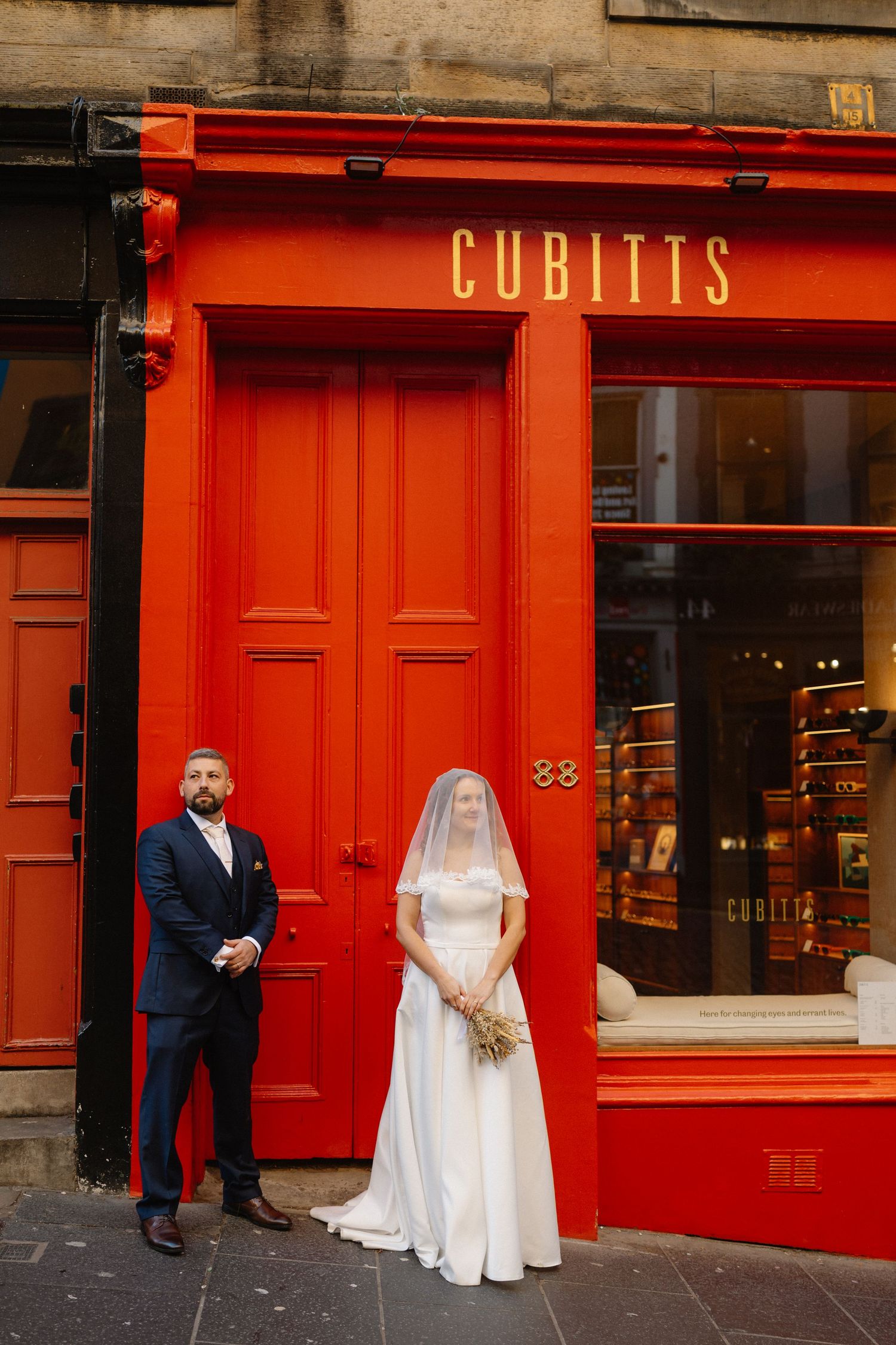 Wedding couple poses in front of bright red storefront with Cubitts signage.