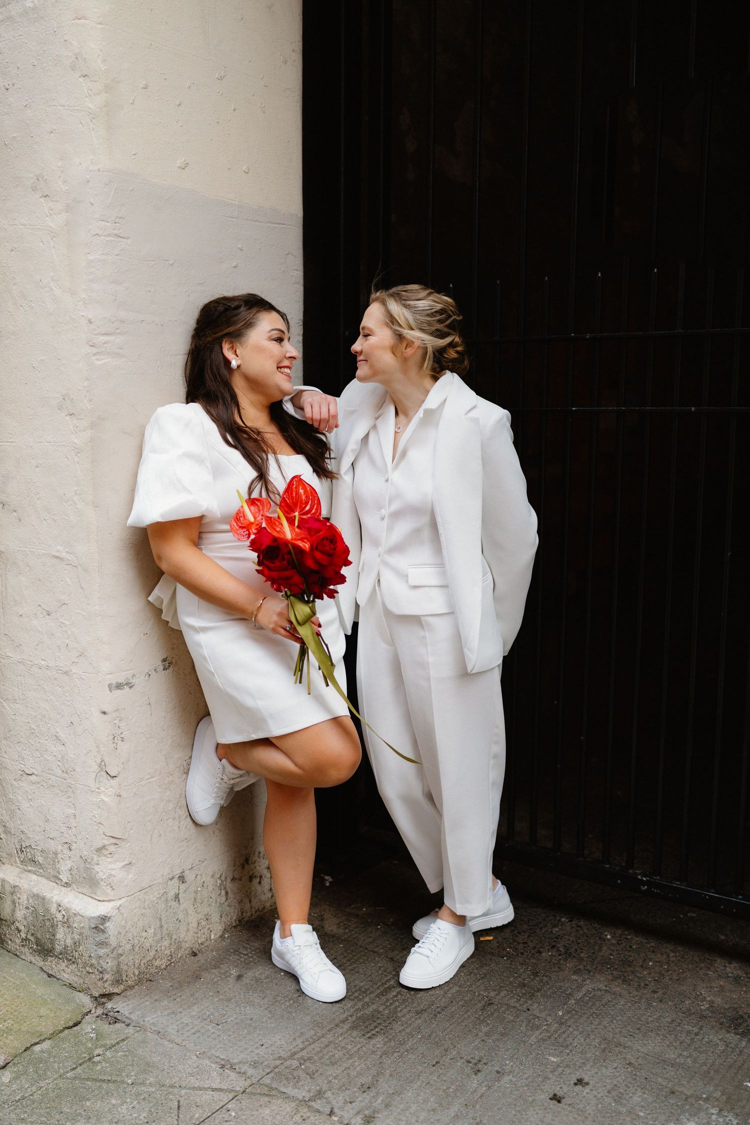 Wedding couple in all white outfits poses against textured wall with red bouquet.