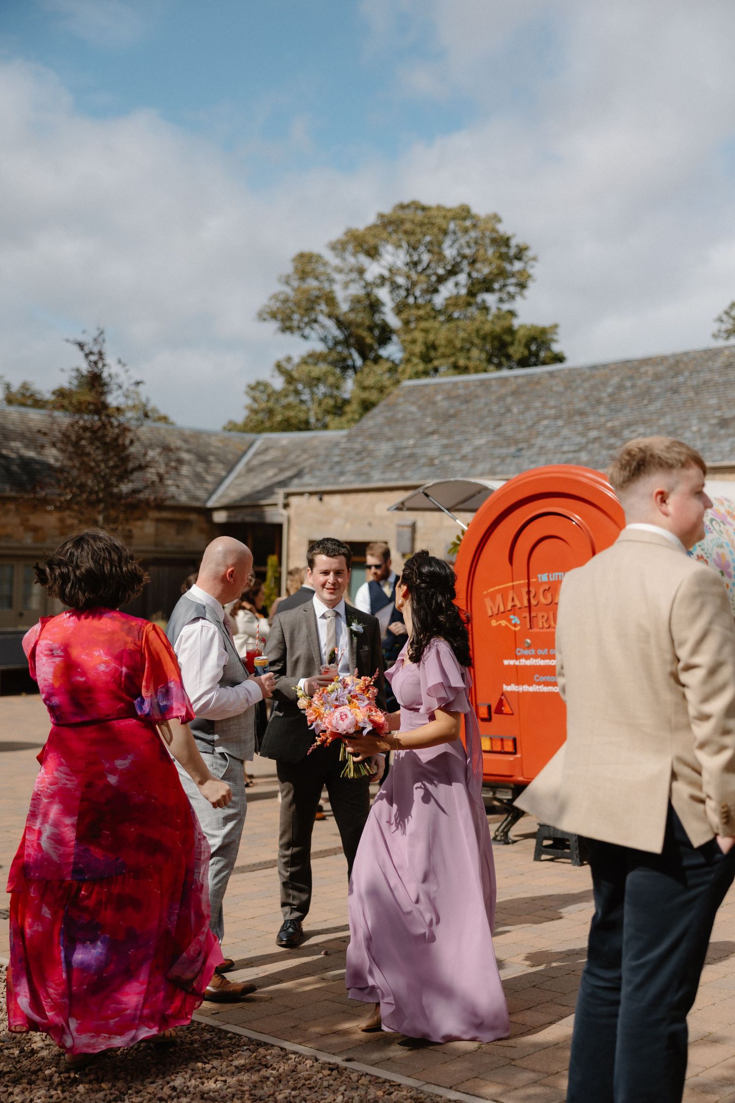 Wedding guests gather outside venue near bright orange structure under cloudy sky.