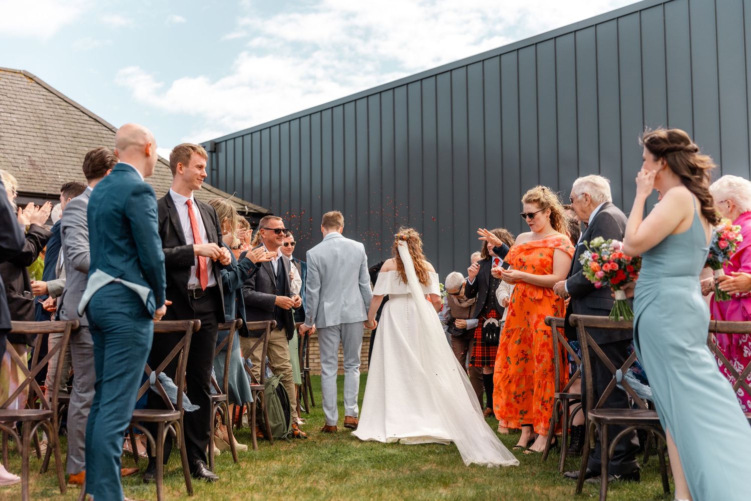 Wedding ceremony guests gather around couple in outdoor setting with industrial backdrop.
