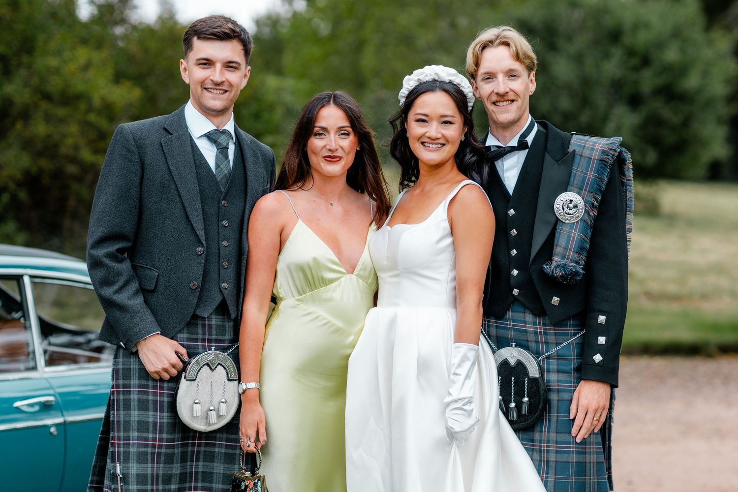 Wedding party poses together outdoors with vintage car and greenery in background.
