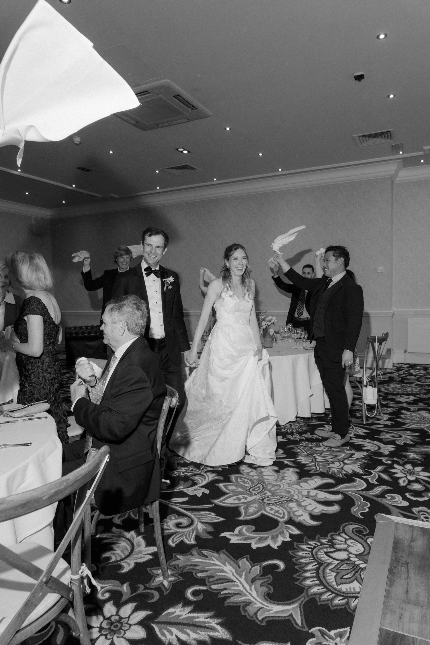 Black and white photo of bride and groom entering wedding reception as guests wave napkins.
