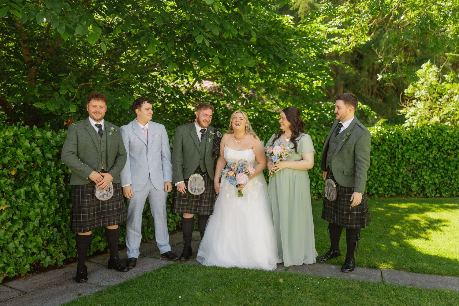 Wedding party posing outdoors in lush garden, bride in white gown with bridesmaids in sage green and groomsmen in tartan kilts.