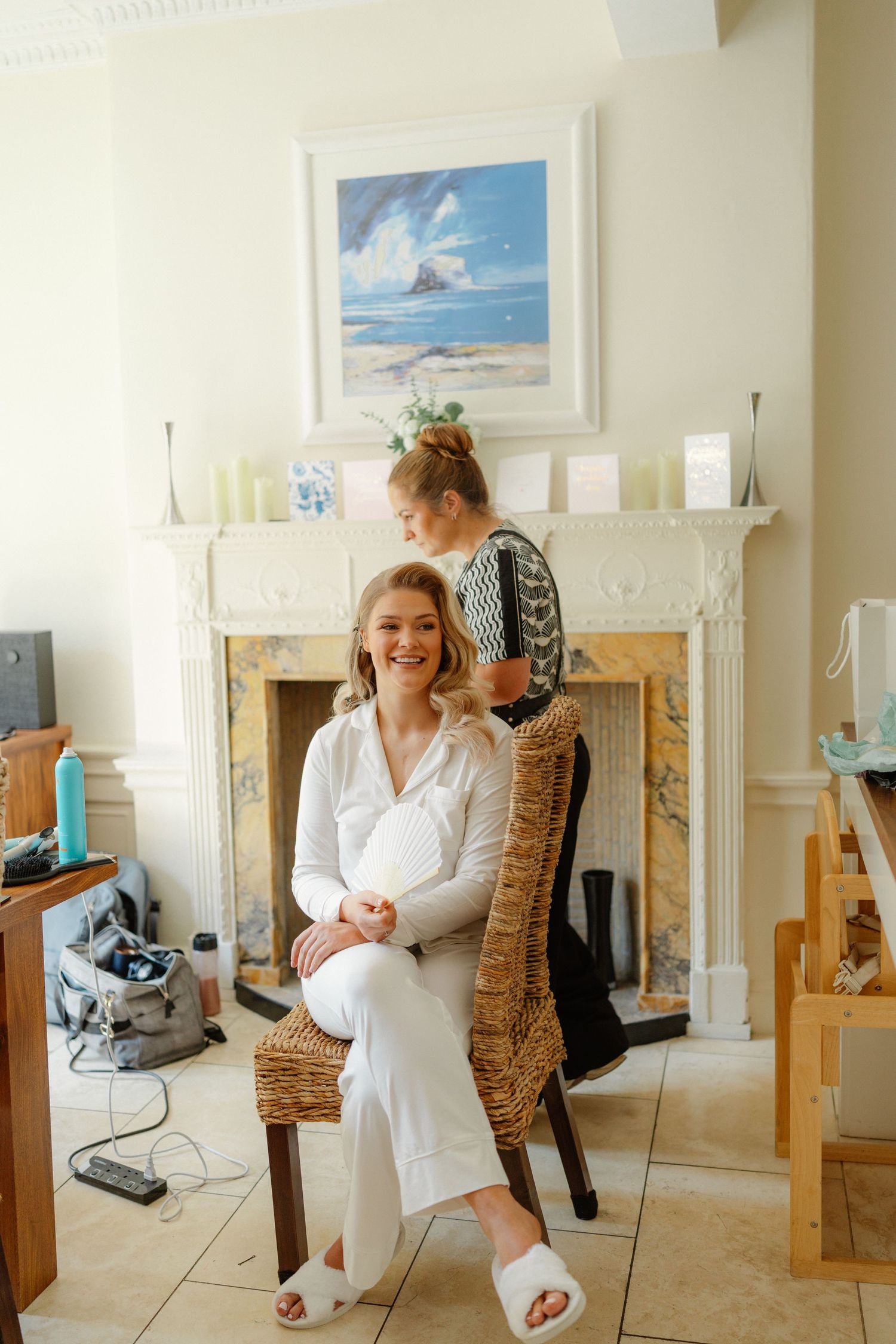 Smiling bride in white outfit sits in wicker chair getting hair done before wedding, elegant room with fireplace backdrop.