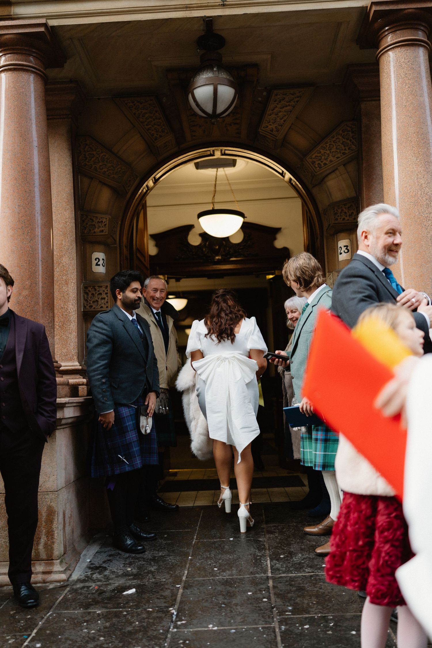 Bride in white dress exits grand arched doorway surrounded by wedding guests in formal attire.