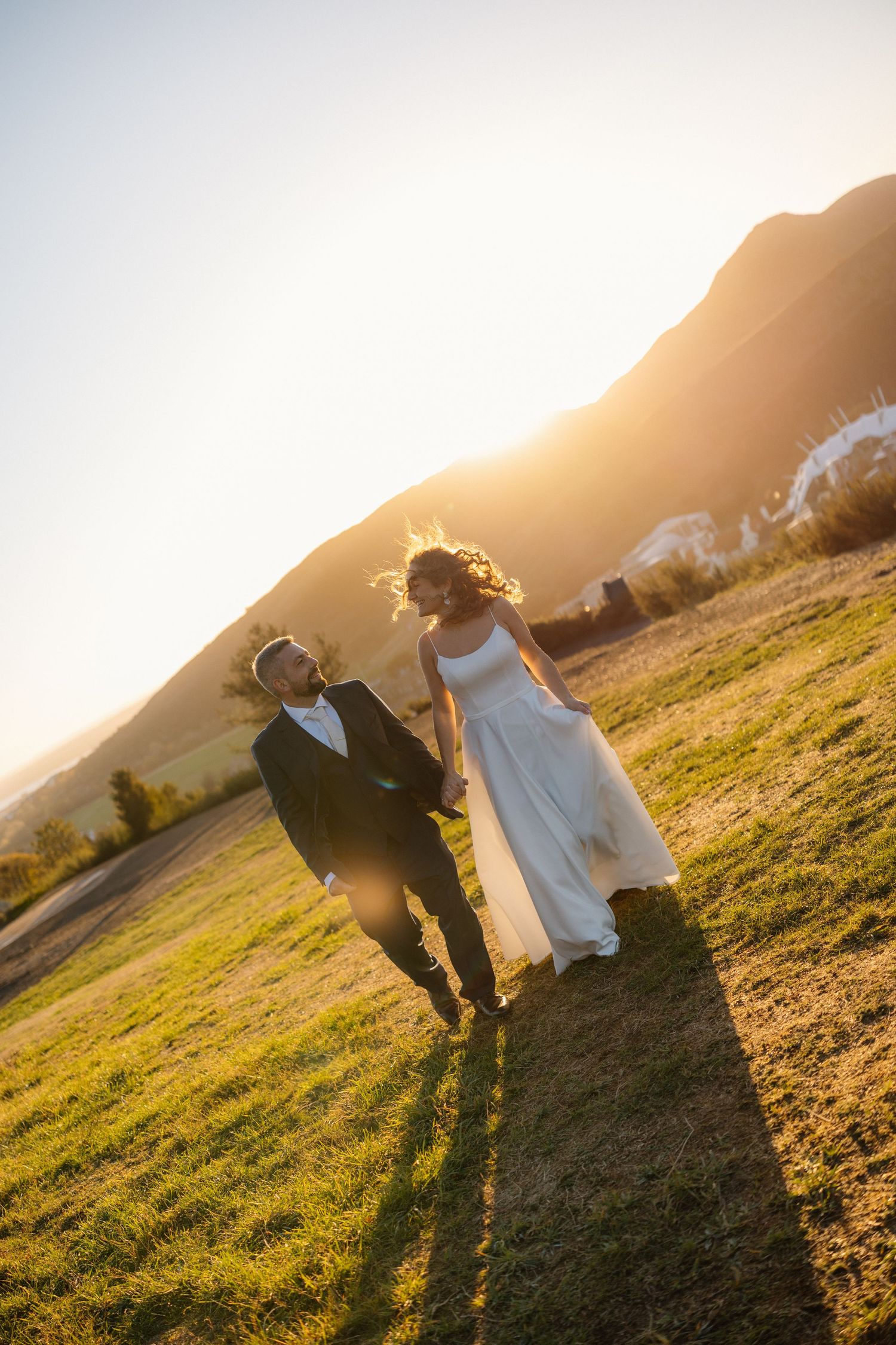 A couple in wedding attire dances together on a grassy hillside during golden hour sunset.
