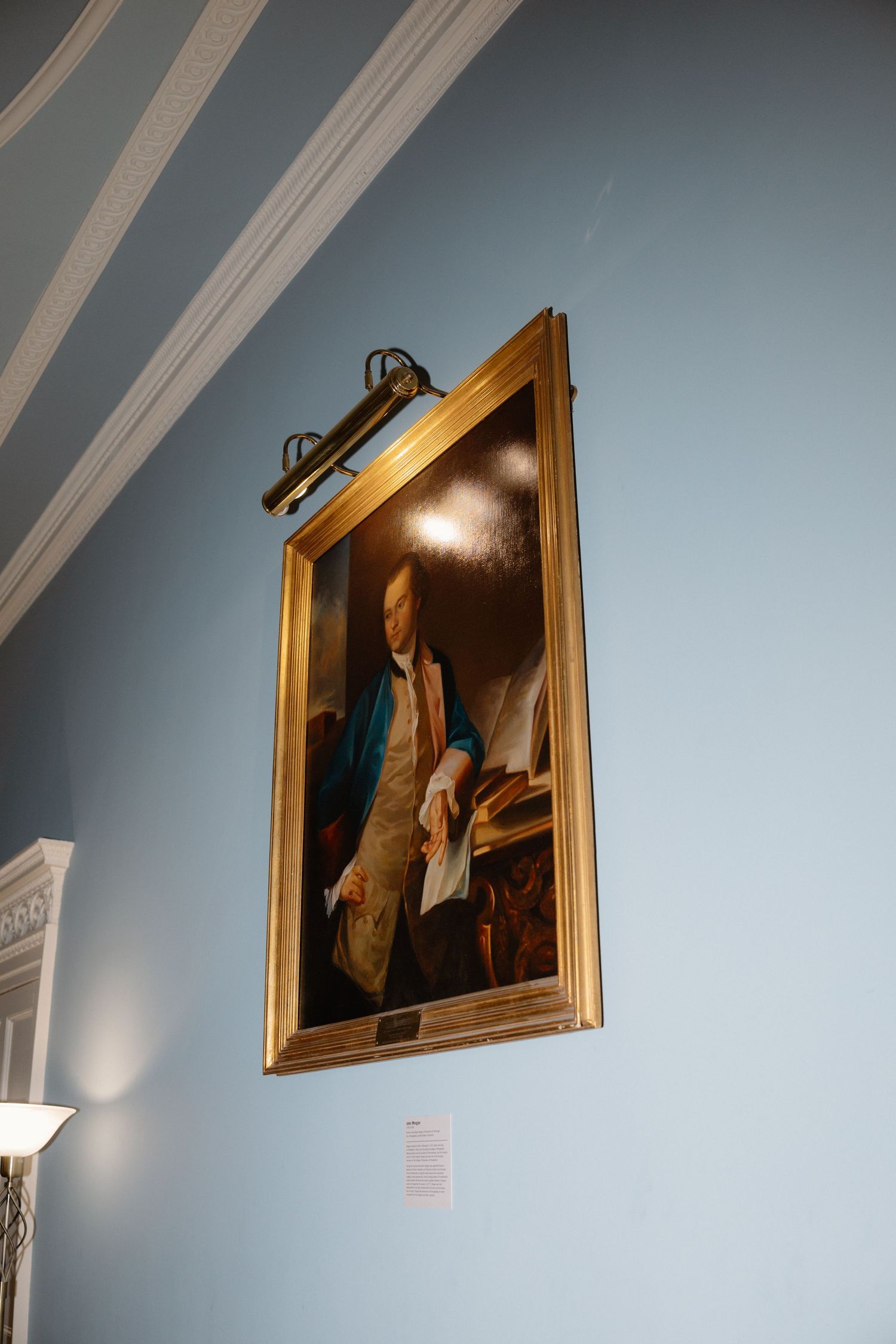 Museum artwork in gold frame mounted on light blue wall with gallery lighting illuminating the classical painting.