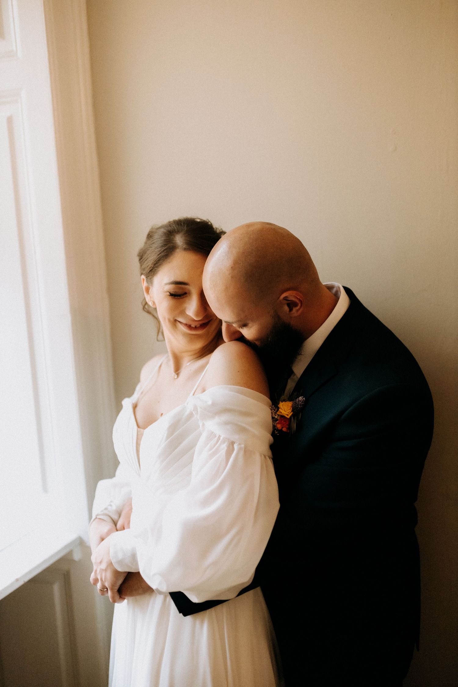 A tender moment between newlyweds in formal attire by a bright window.