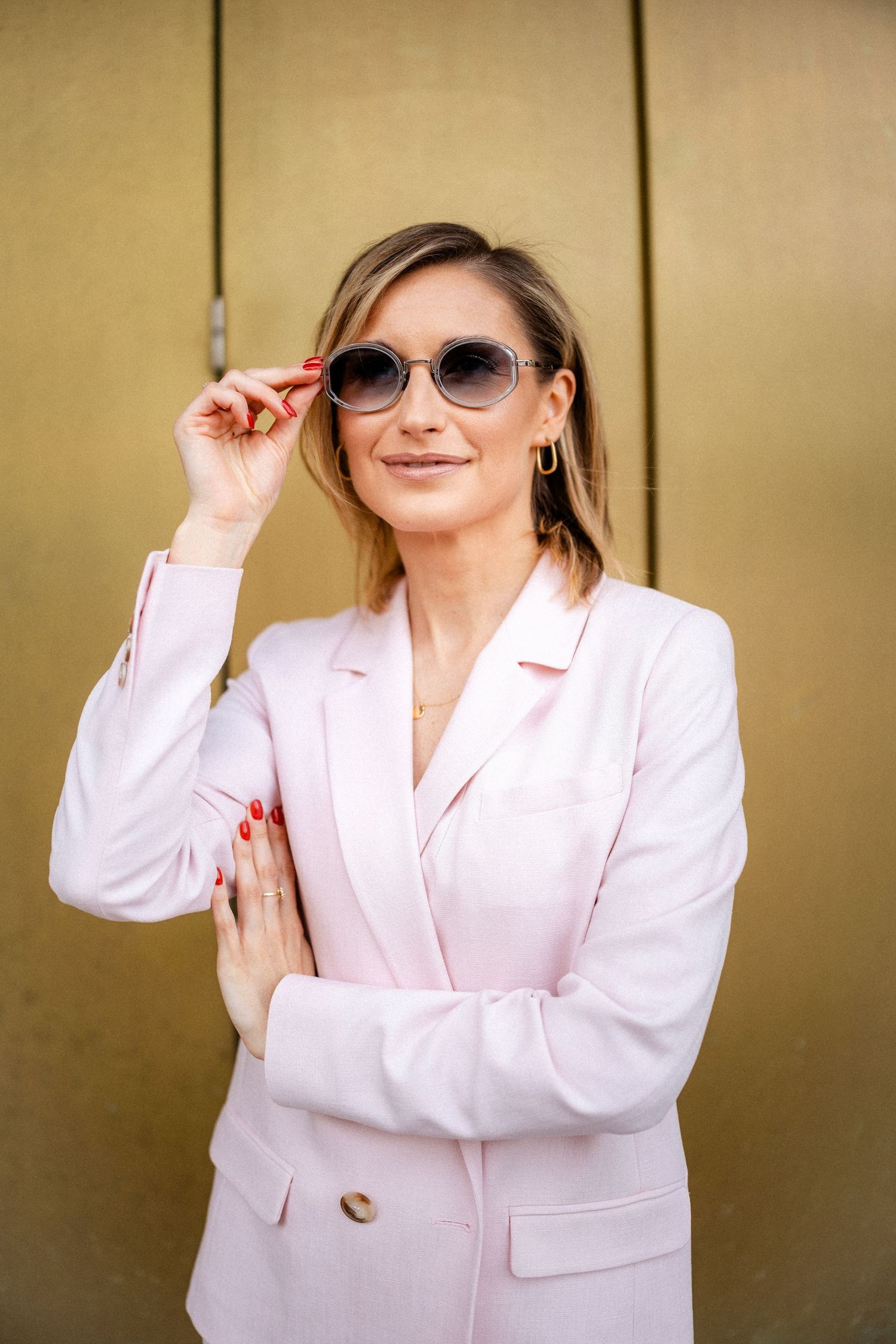 Portrait of someone in pink blazer adjusting sunglasses against gold background.
