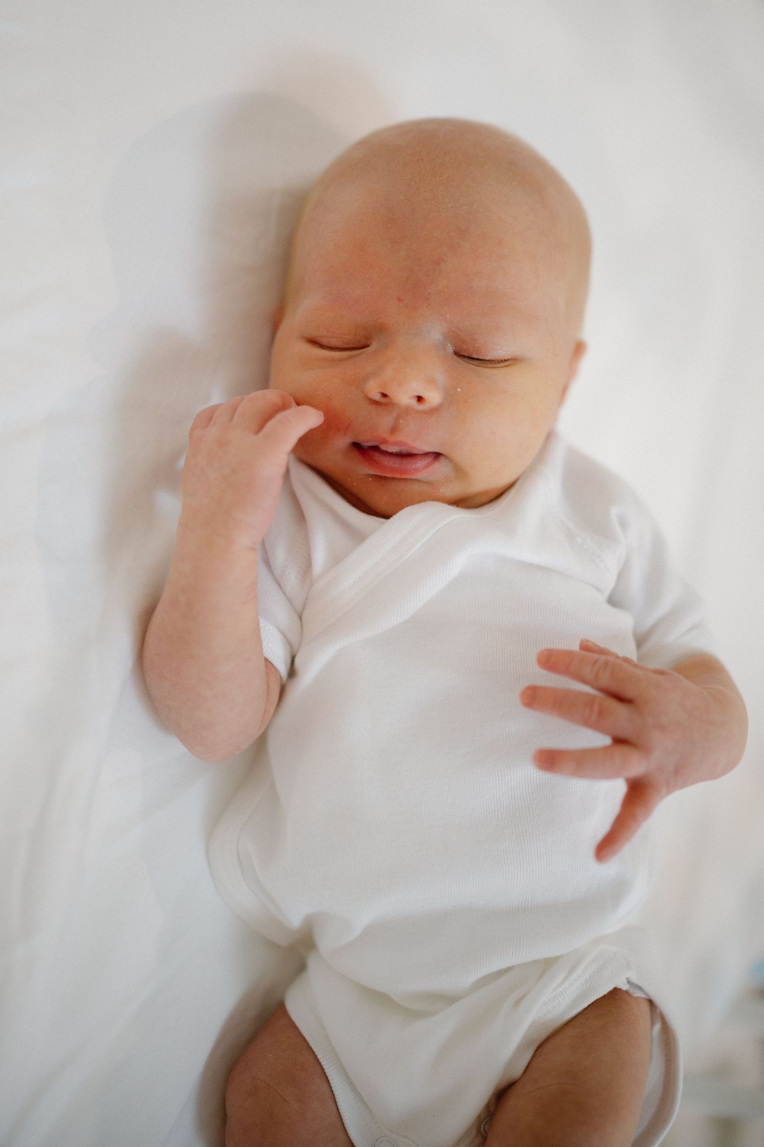 Newborn baby sleeping peacefully in white onesie on soft bedding.