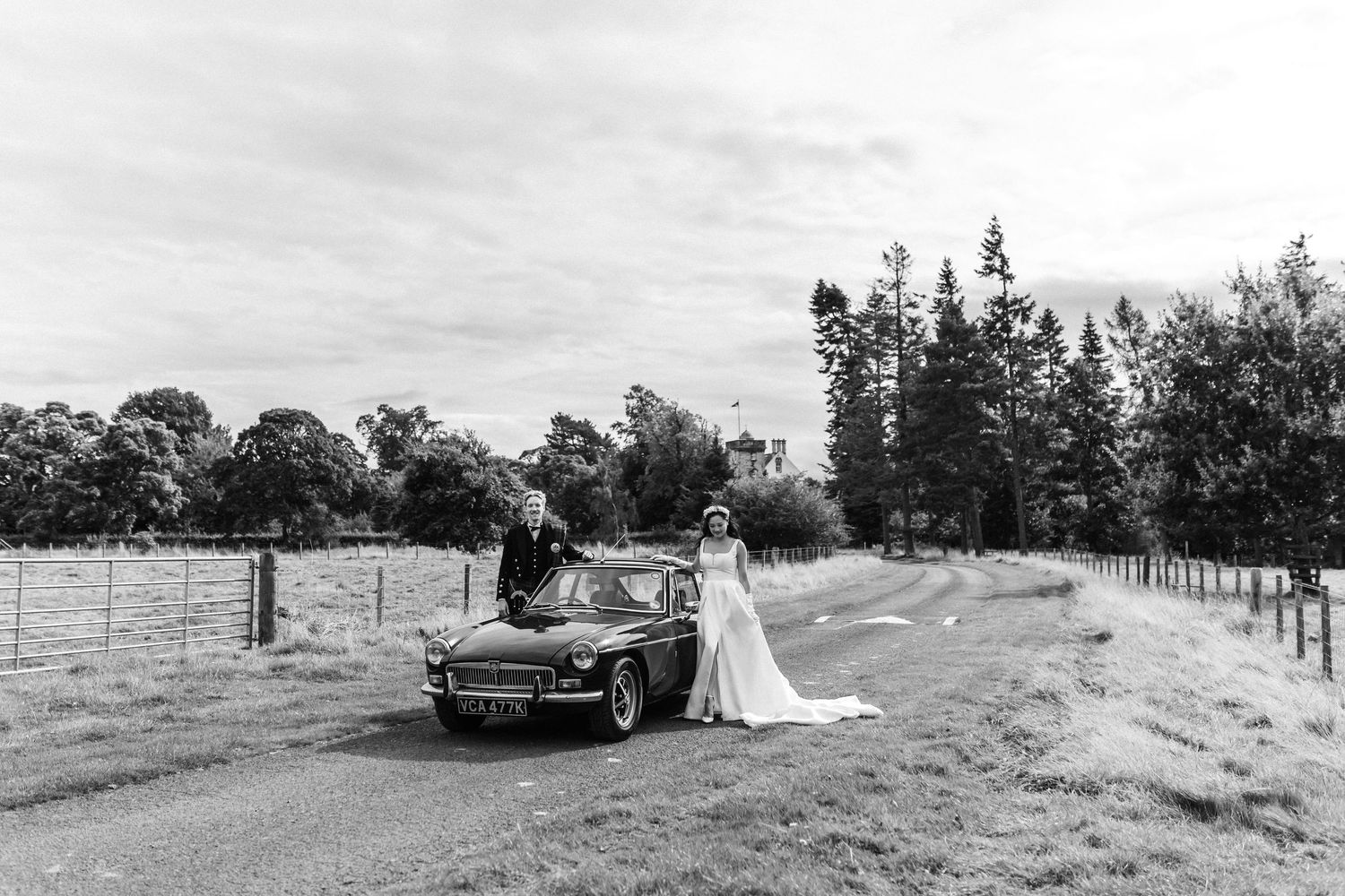 Wedding couple posing with classic car in rustic outdoor setting.