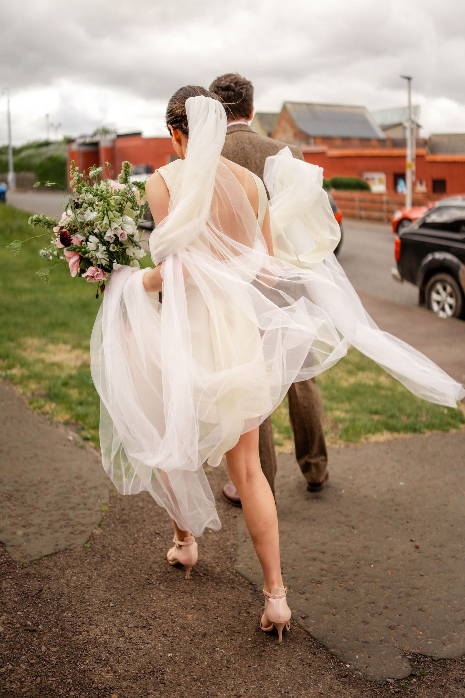 The bride's dress flows dramatically as she walks through a parking area.