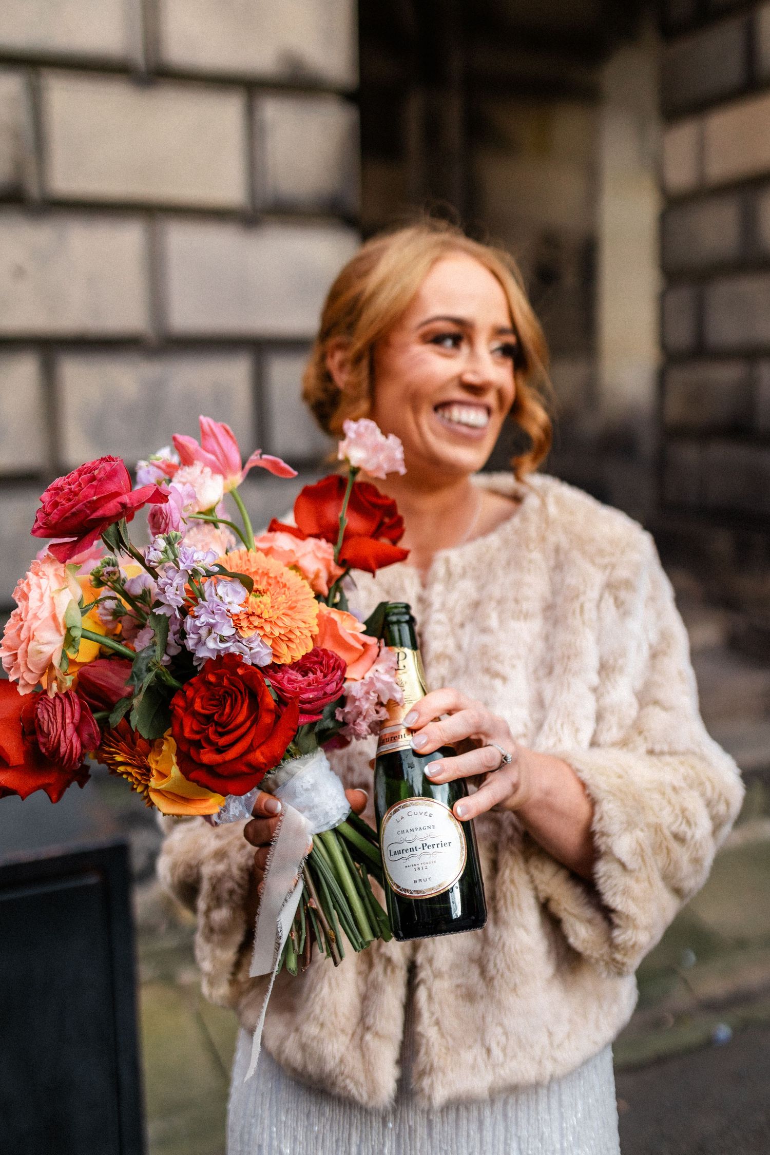 Bride in fur wrap holds vibrant bouquet of red and pink flowers against stone wall.