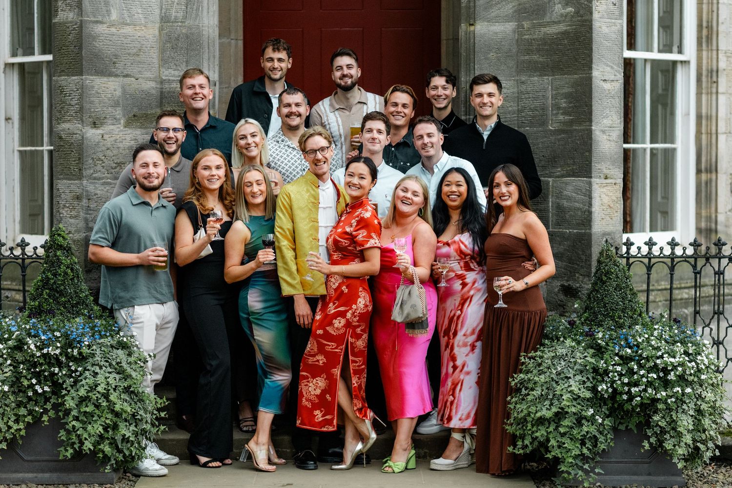Wedding party and guests pose for a group photo outside a historic stone building.