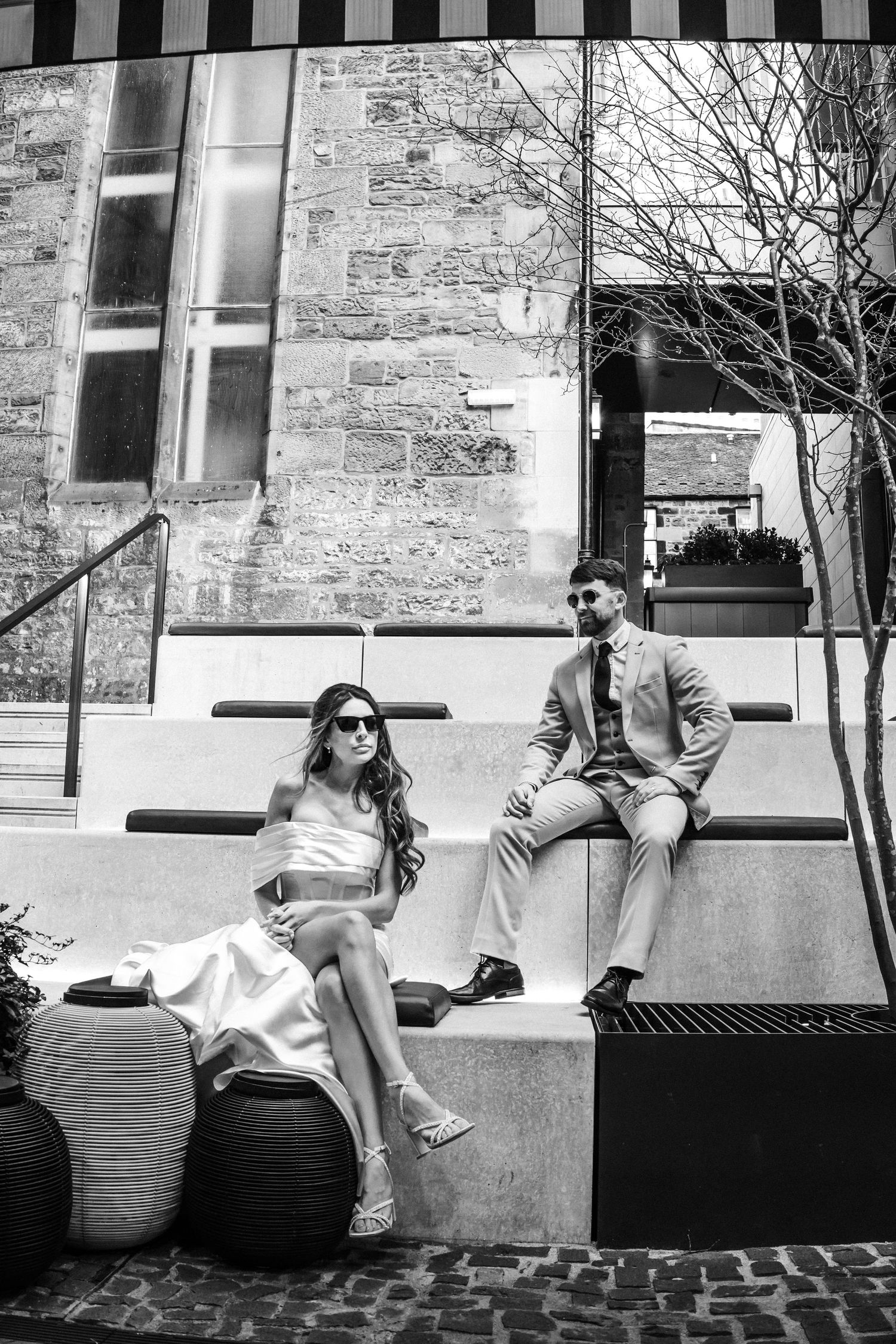 Couple poses playfully on stone benches in black and white photography.