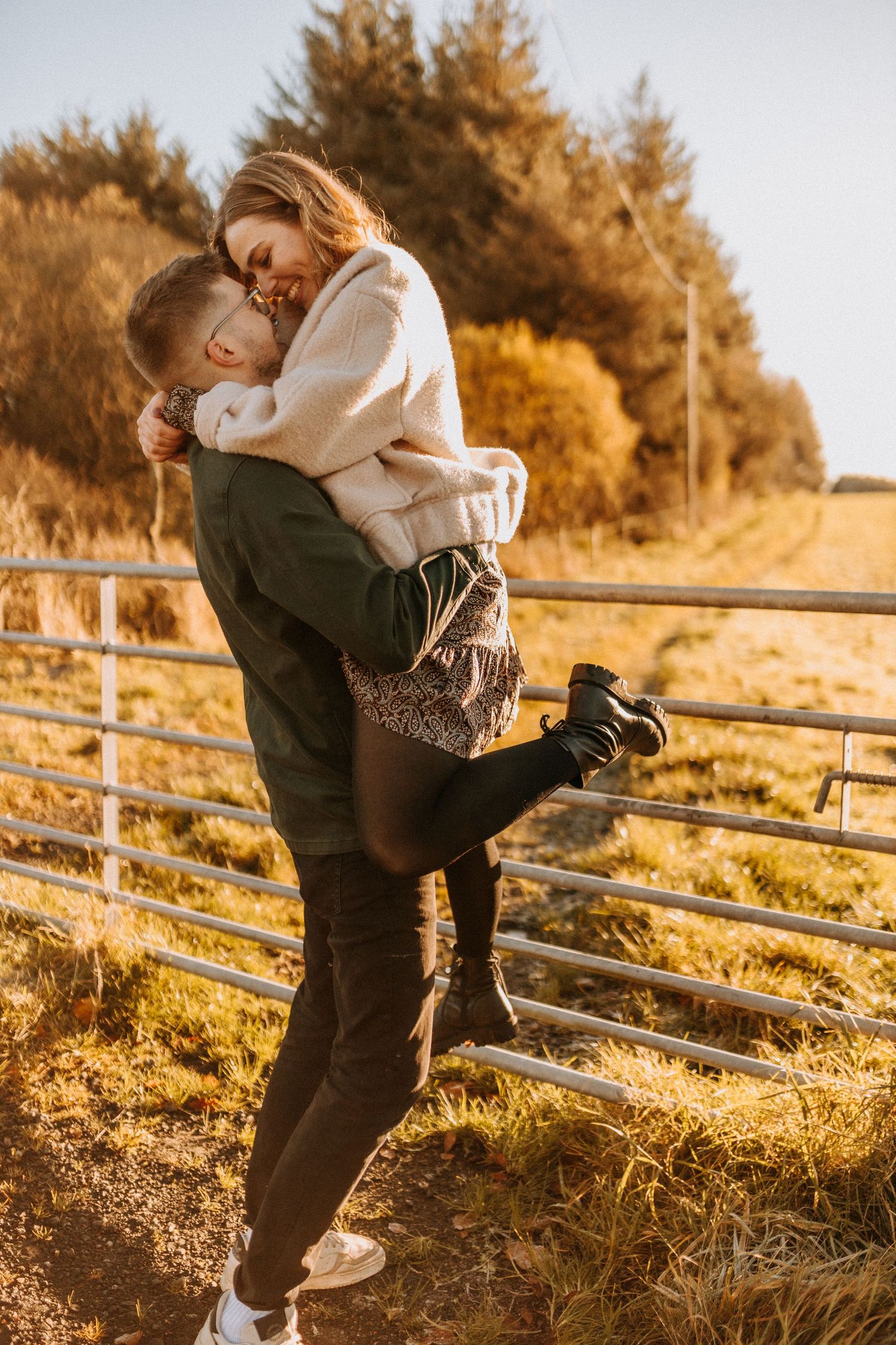 Sunset engagement photos by a rustic wooden fence in golden hour lighting.