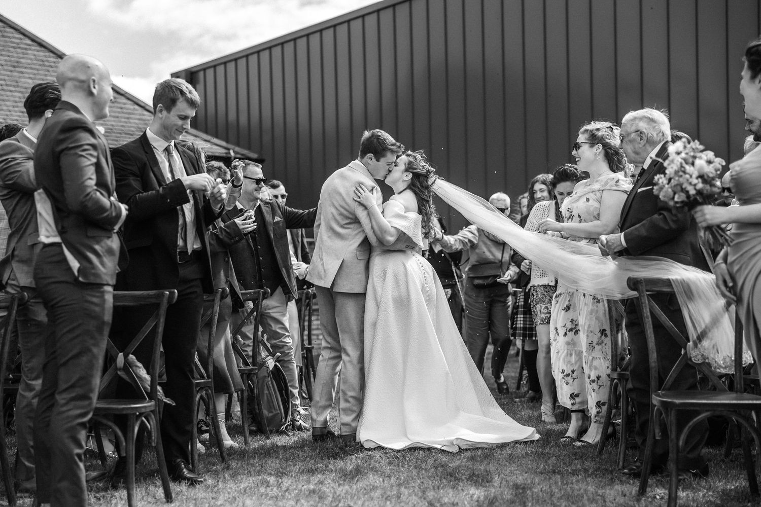Black and white photo of wedding guests seated outdoors with a metal building in the background.
