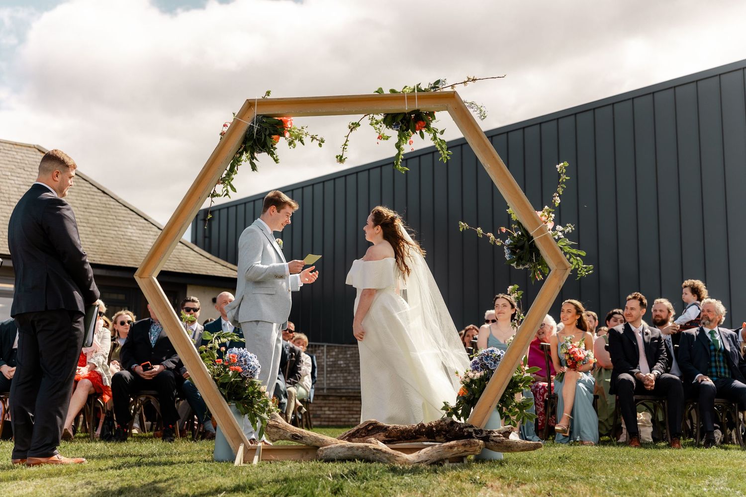 Wedding ceremony under wooden arch with guests celebrating in outdoor venue setting.